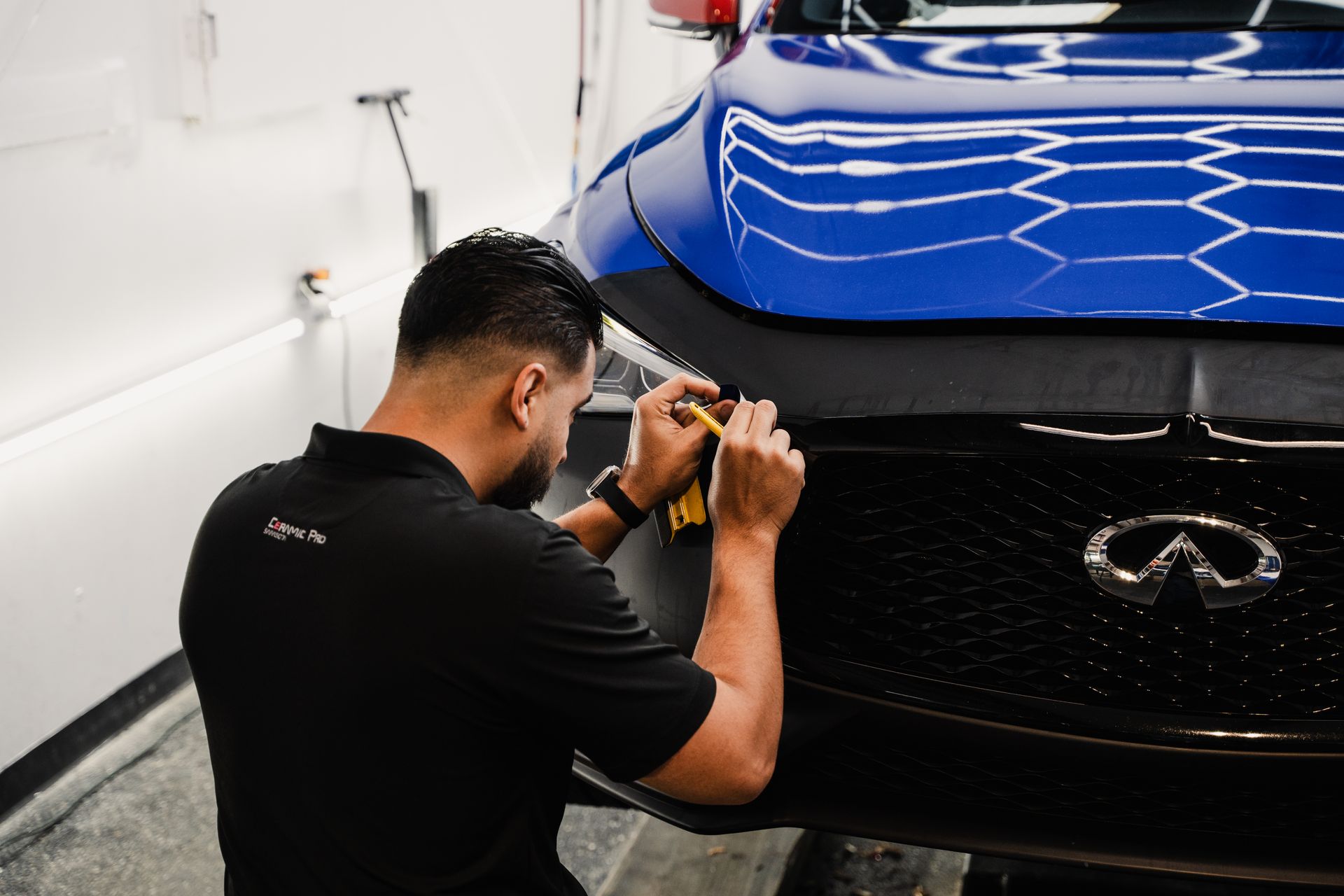 Technician polishing the front of a blue Infiniti car in an auto detailing shop