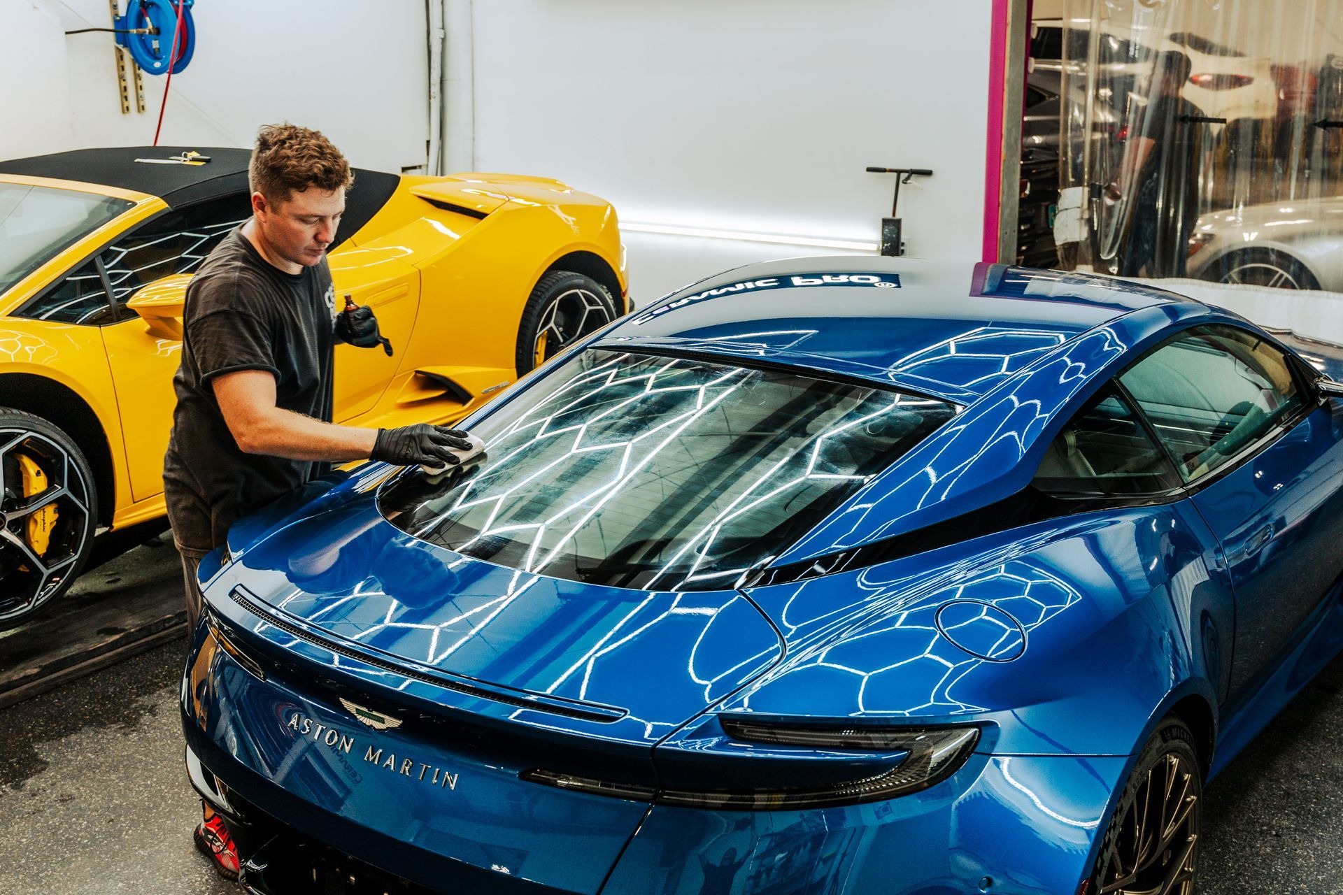 Detailer polishing a blue sports car in a garage beside a yellow supercar.