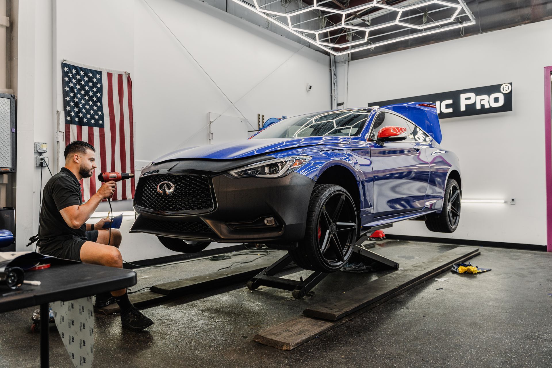 Blue sports SUV on a lift in a garage, with a technician kneeling nearby and an American flag on the wall.