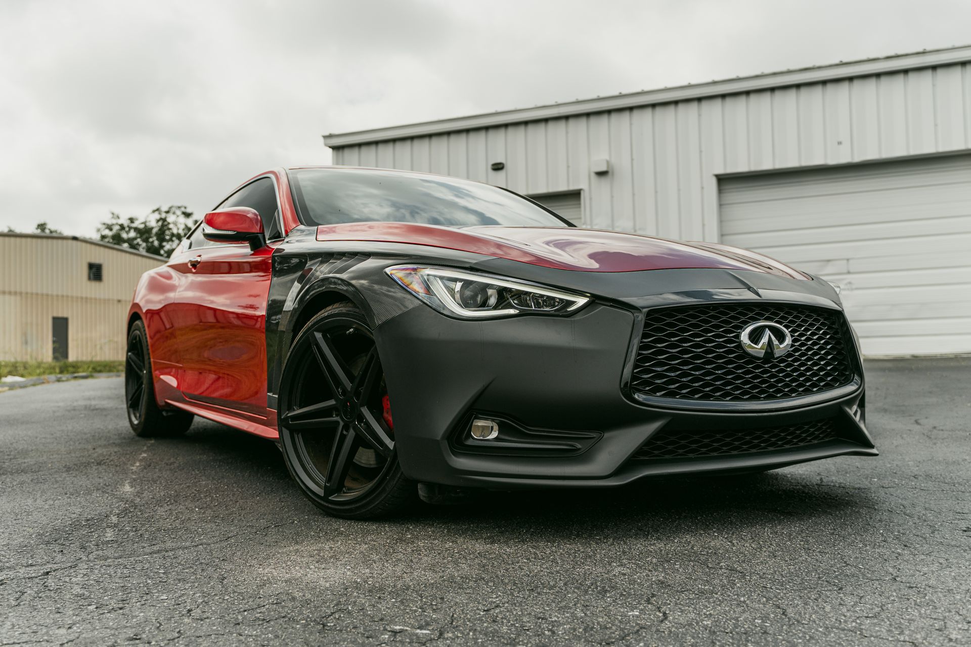 Red Infiniti sports sedan parked on wet pavement beside a white industrial building.