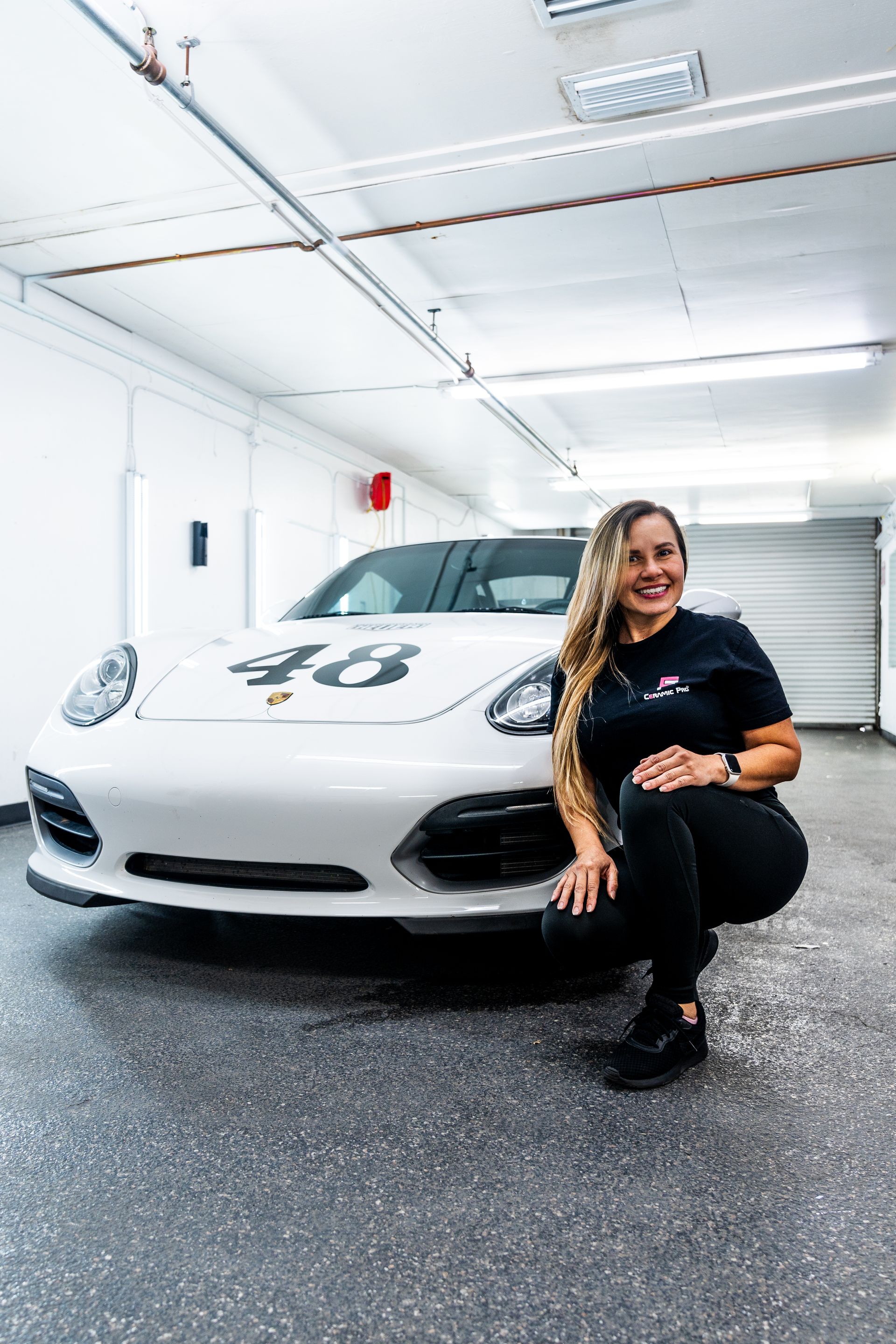 Woman crouching beside a white sports car in a bright garage, smiling at the camera.