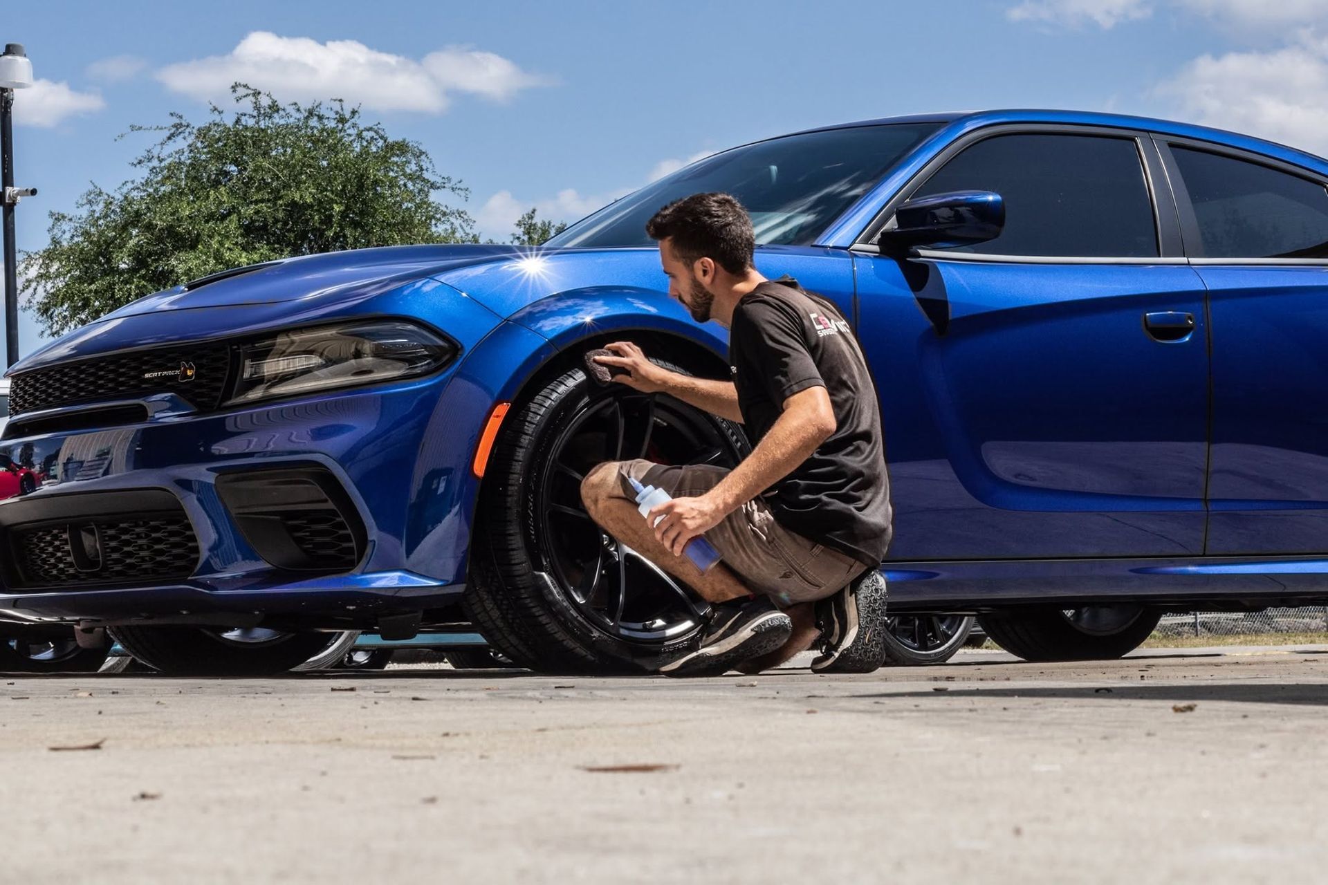 Man crouching by a blue sports sedan, cleaning or inspecting the front wheel outdoors
