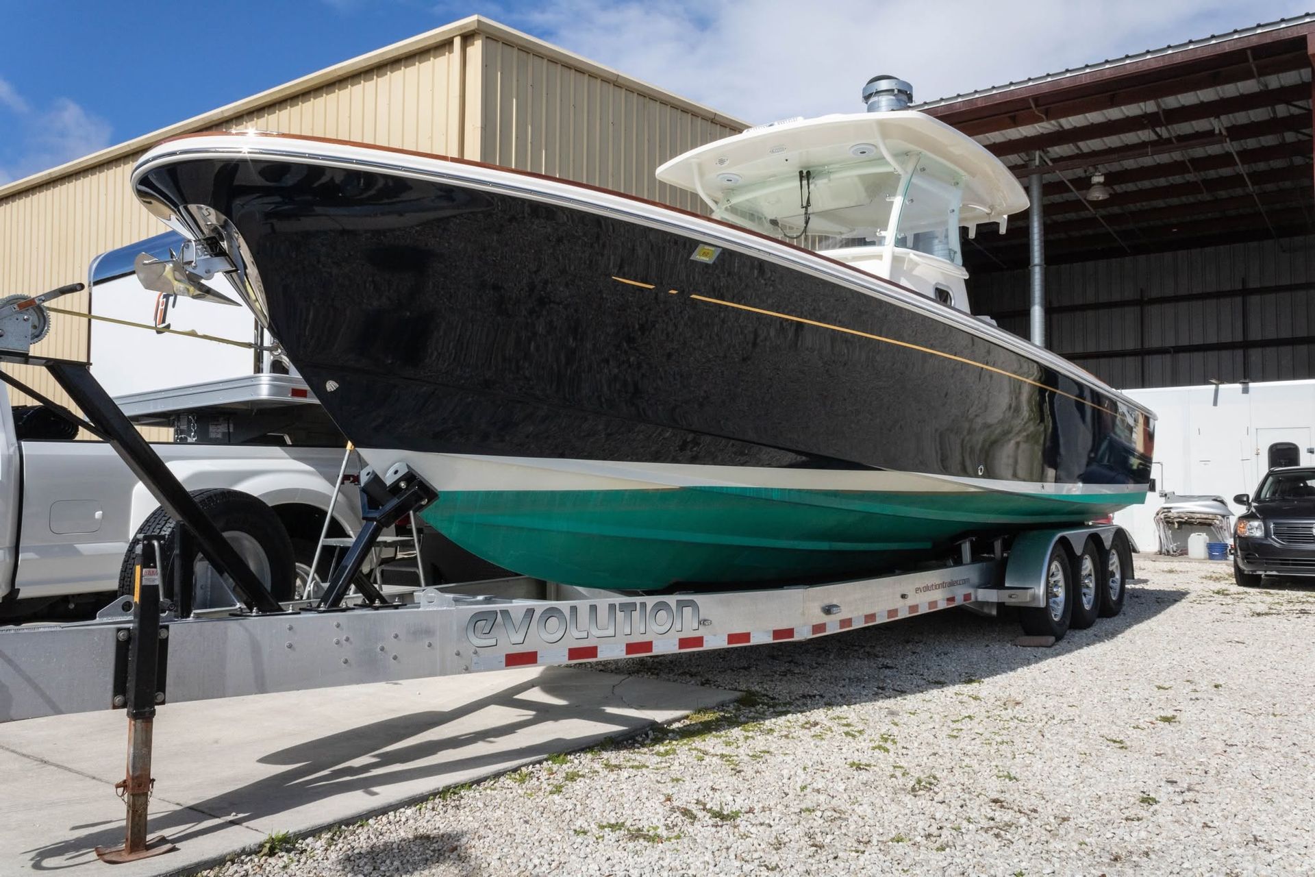 Large black-and-teal speedboat on a trailer in a gravel lot beside a warehouse