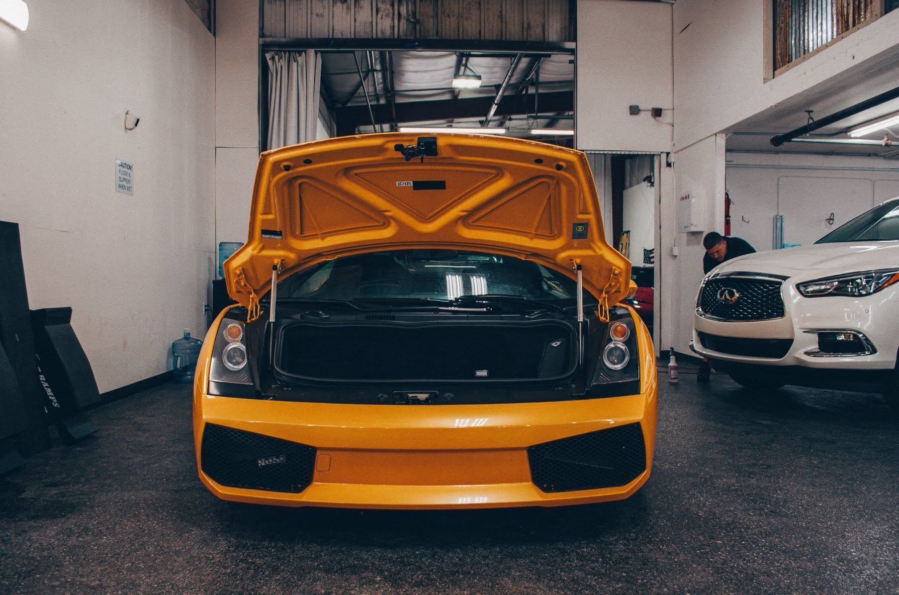 Yellow sports car with hood open in a garage, with a white car parked beside it.