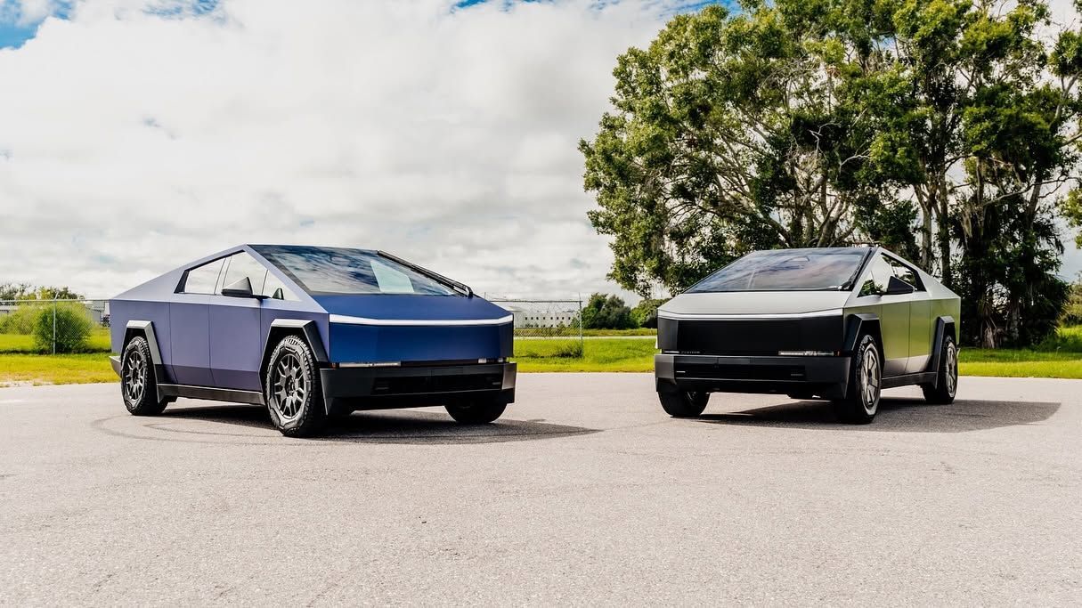 Two sleek electric pickup trucks, one blue and one black, parked on a road by a tree