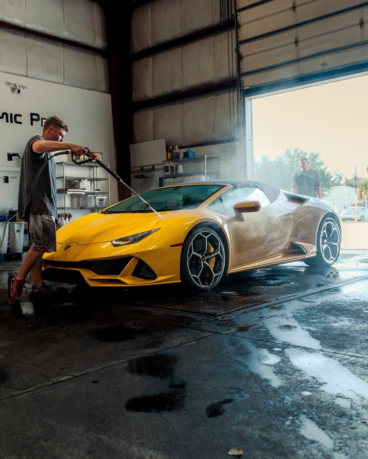 Yellow sports car being washed in a garage by a worker, with water spray and wet floor.