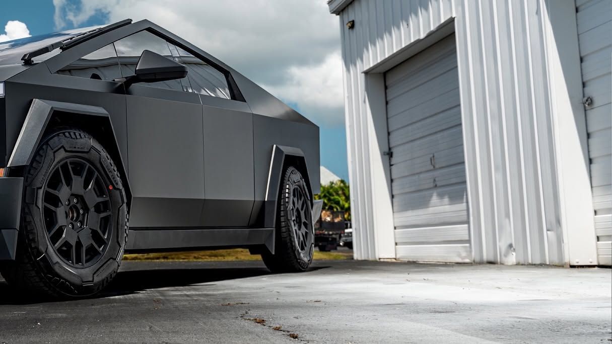 Matte black futuristic pickup truck parked beside a white garage under cloudy skies