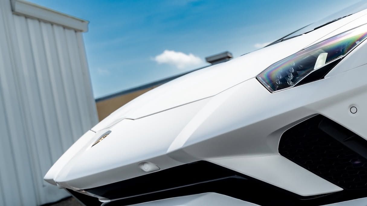 Close-up of a white sports car’s front end beside a building under a blue sky