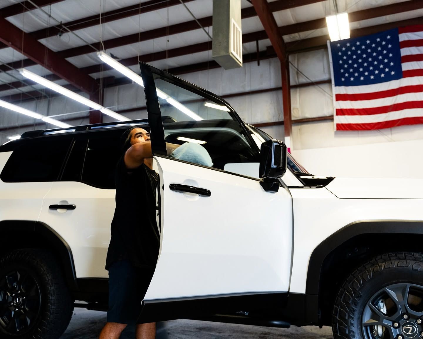 Person holding a vehicle door open in a garage, with a white SUV and an American flag in view