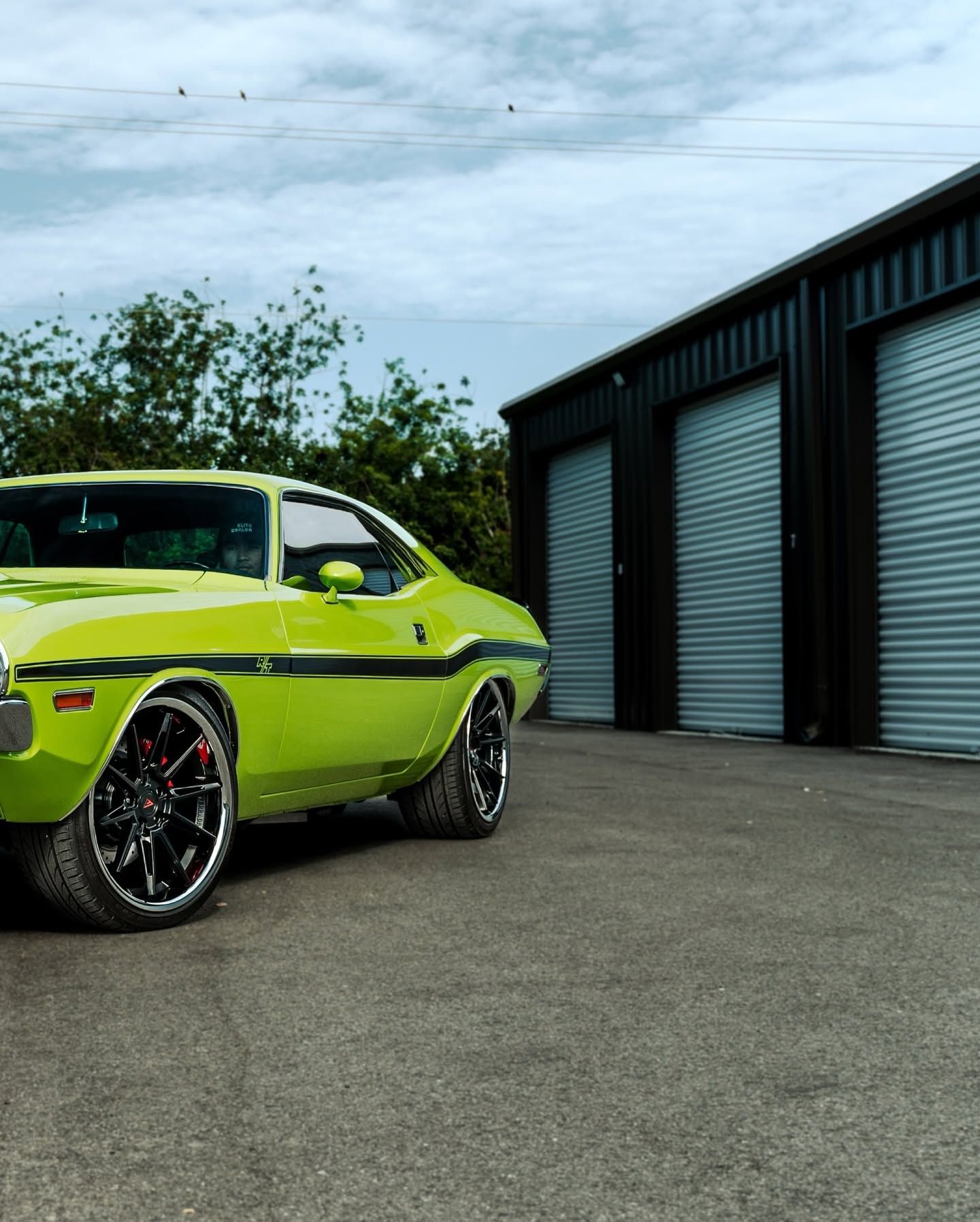 Lime green classic car parked beside black industrial garage doors under a cloudy sky.