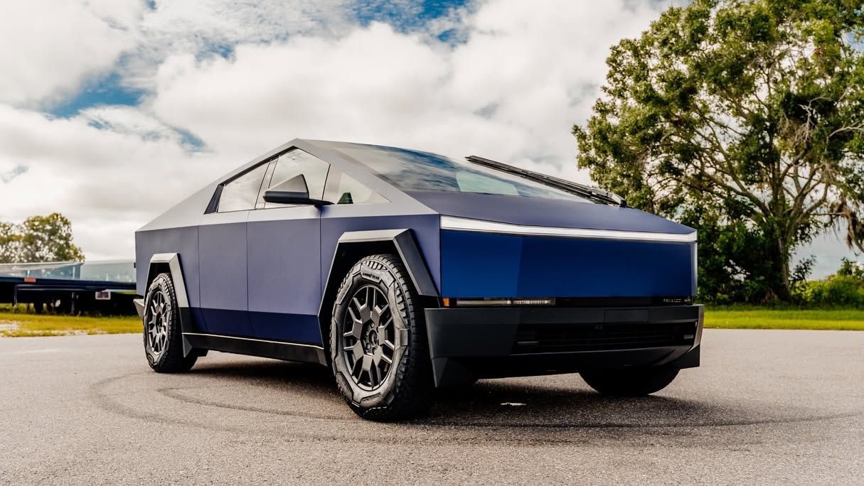 Blue Tesla Cybertruck parked on a road under a cloudy sky, seen from the front-left side