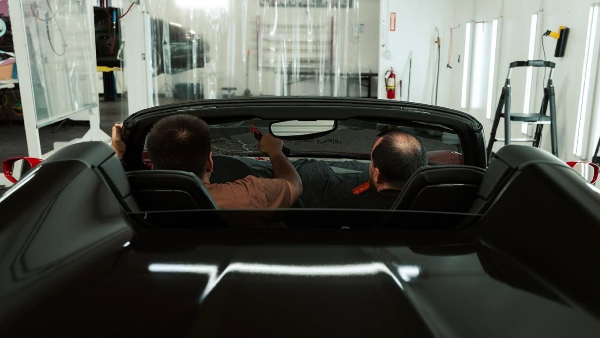 Two people sit in a black convertible inside a bright showroom, viewed from the back seat.