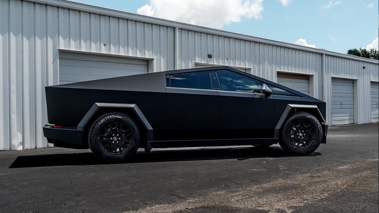 Black Tesla Cybertruck parked beside industrial warehouses on an overcast day
