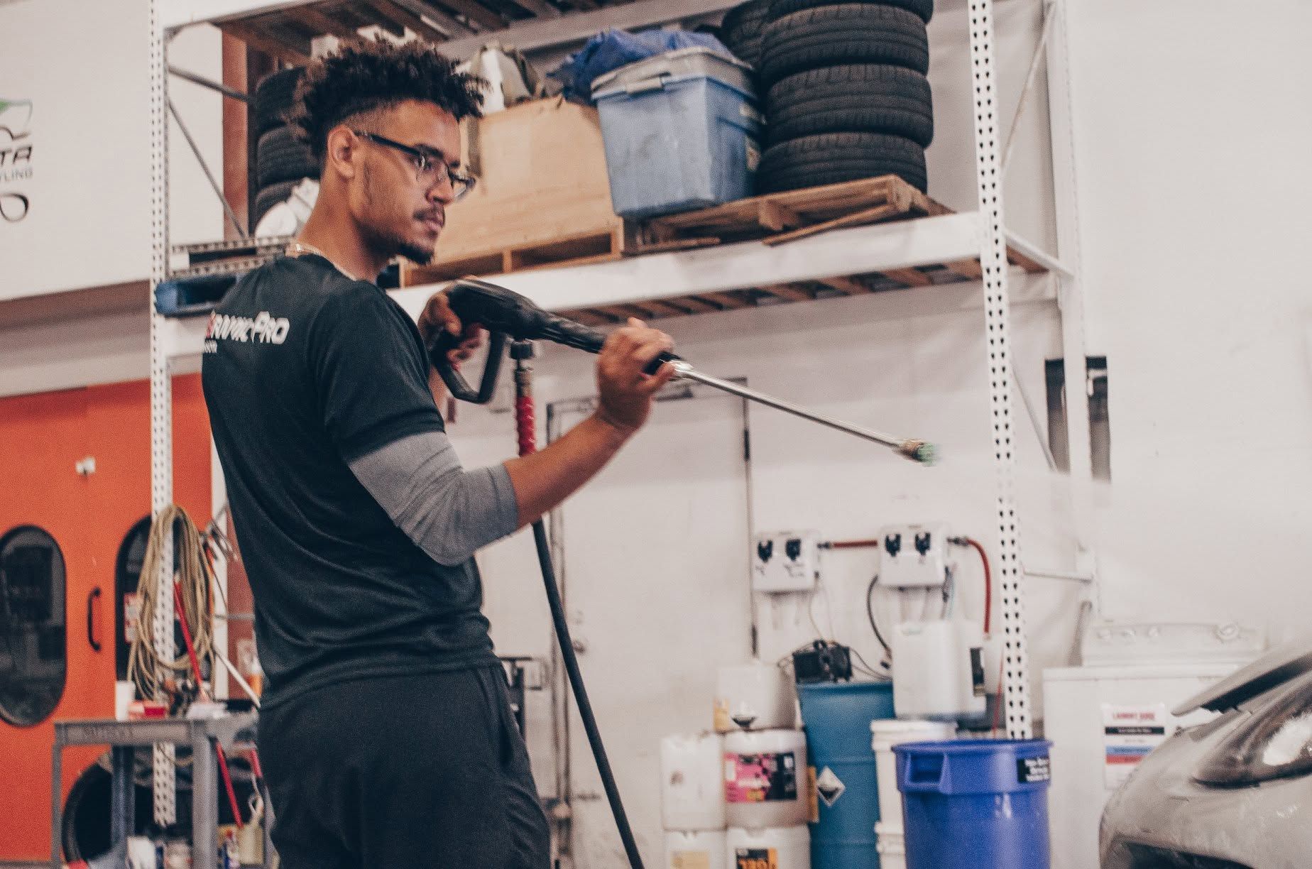Worker using a pressure washer in a tire shop, spraying a car in a garage