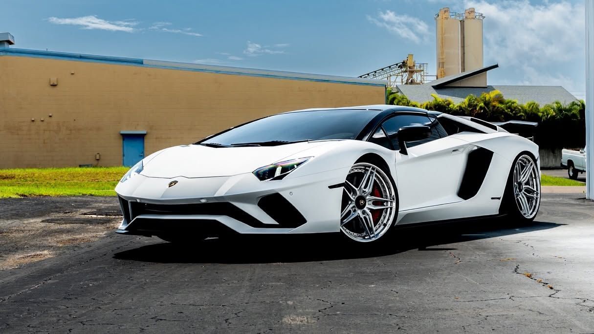 White Lamborghini sports car parked on wet pavement beside a tan building under a blue sky