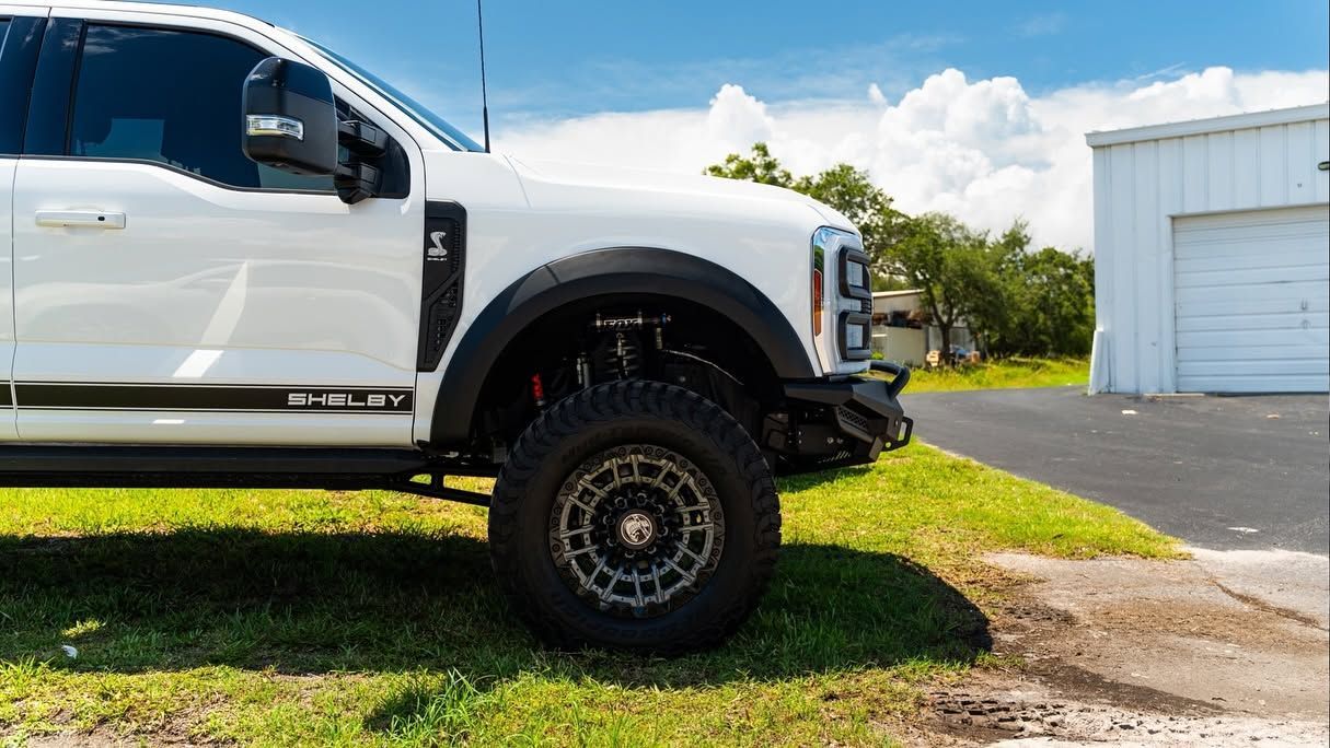 Lifted white pickup truck parked on grass beside a paved lot and white building under a blue sky
