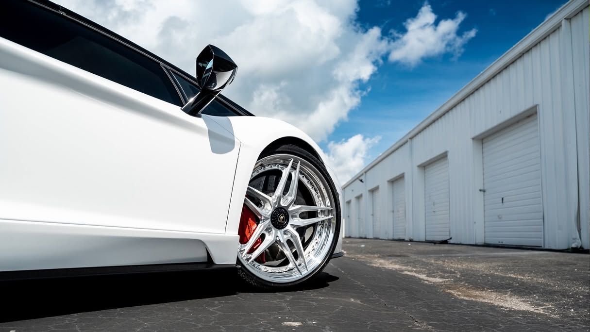 Low-angle view of a white sports car’s chrome wheel parked beside white warehouse buildings under a blue sky