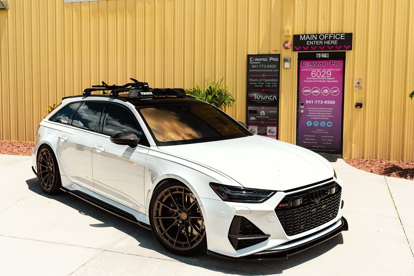 White modified station wagon with black roof and bronze wheels parked by a yellow wall and signboard