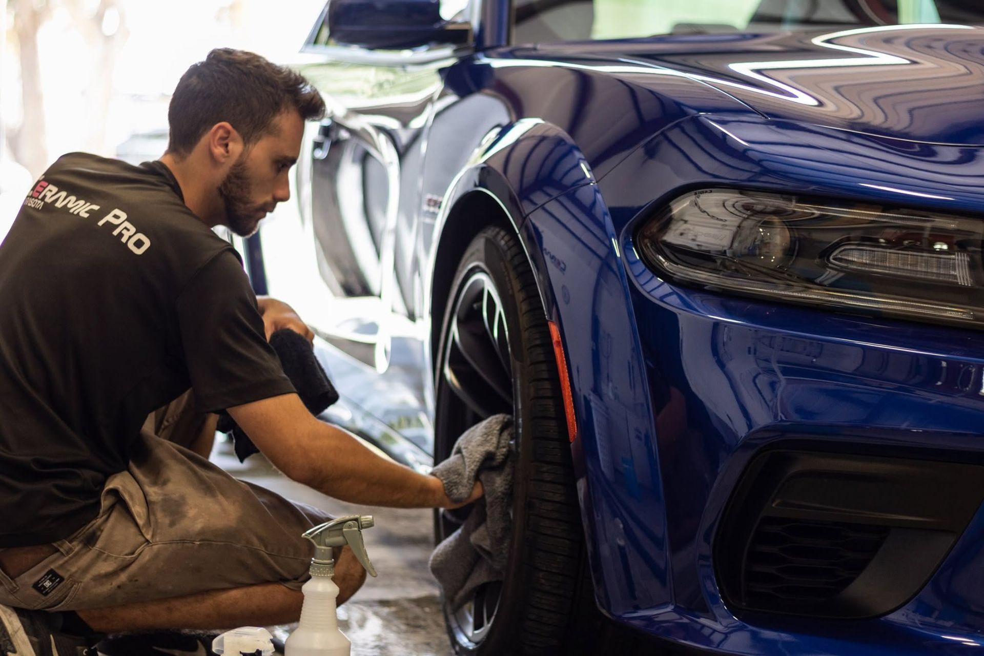 Mechanic kneeling beside a blue sports car, working on the front wheel in a garage.