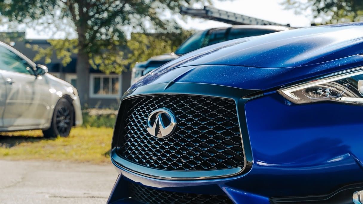 Close-up of a blue Acura grille and emblem, with a silver car blurred in the background.