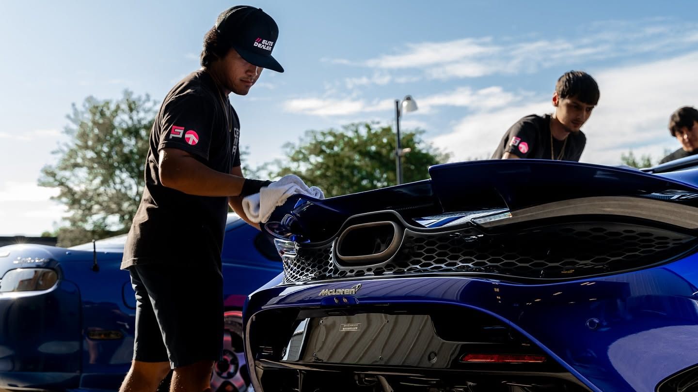 Two people inspect the front of a blue sports car outdoors under a bright sky