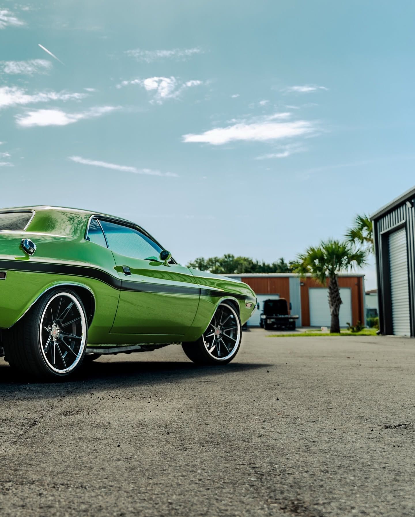 Green classic muscle car parked on pavement beside garages under a blue sky.