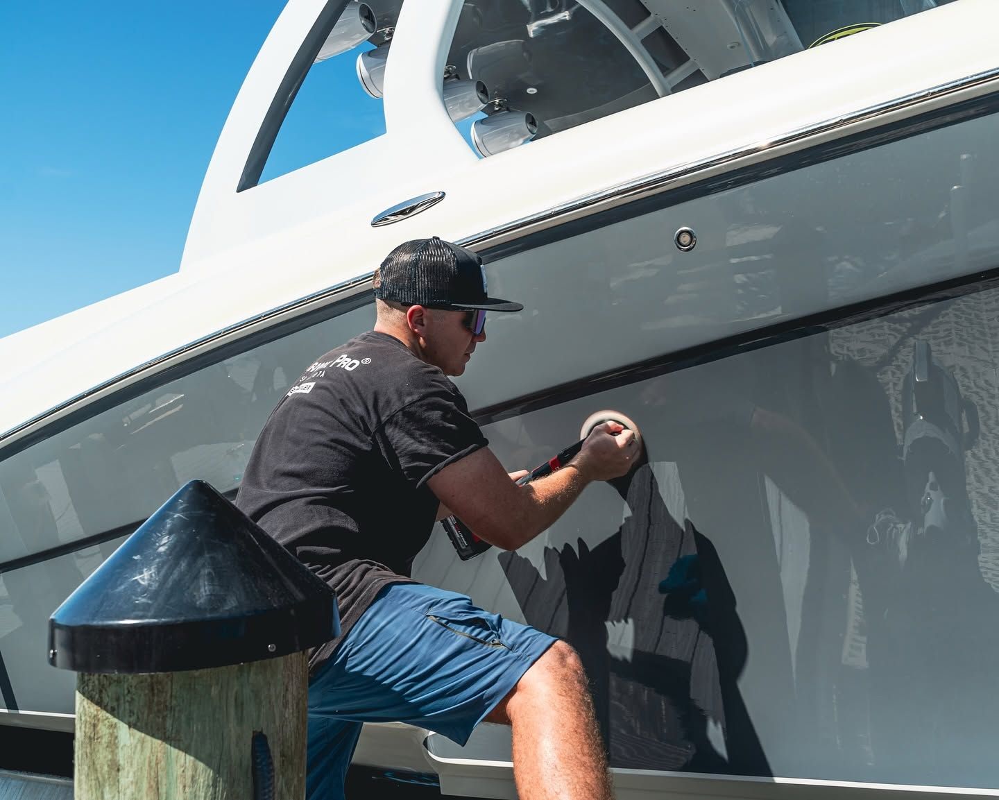 Man scrubbing the side of a white boat while standing on a dock under a clear blue sky