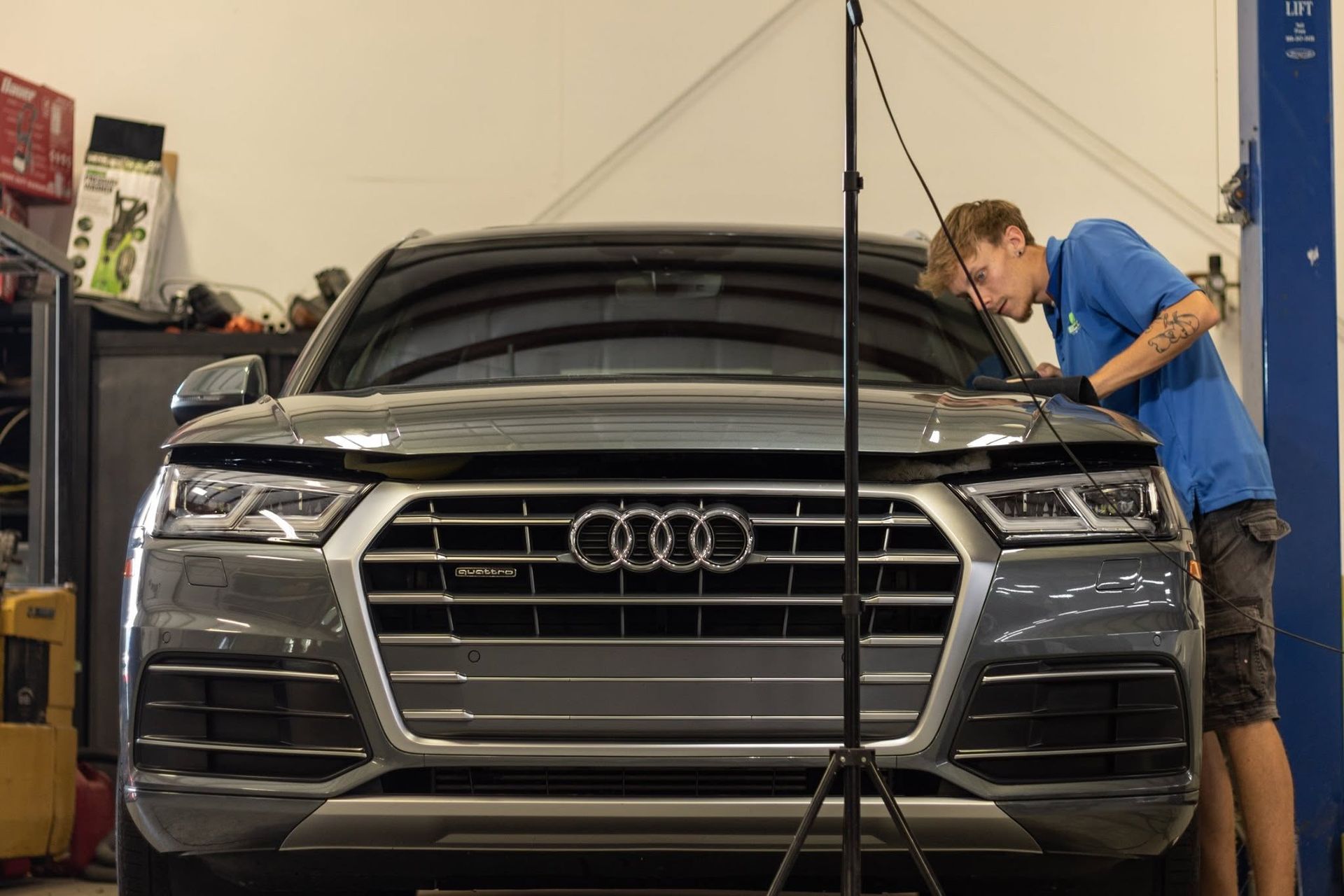 Man working on the front of a silver Audi SUV in a garage, with a lift hood prop visible.