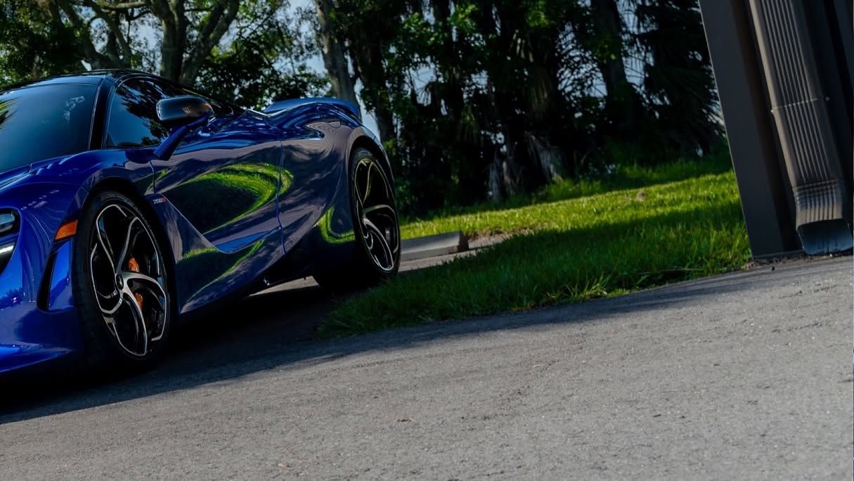 Blue sports car parked on a driveway beside grass and trees, viewed at an angle.