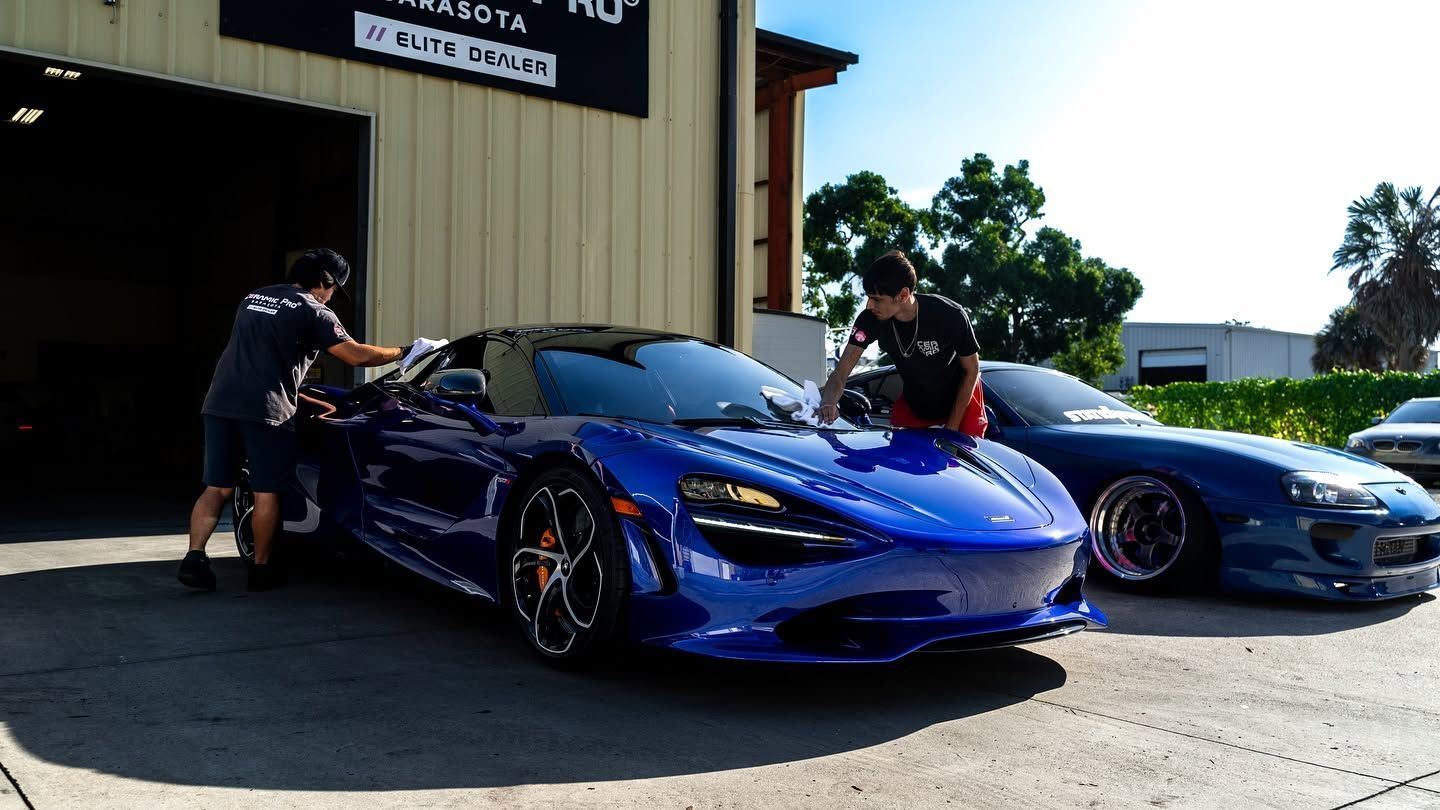 Two men standing by two blue sports cars outside a garage in bright sunlight