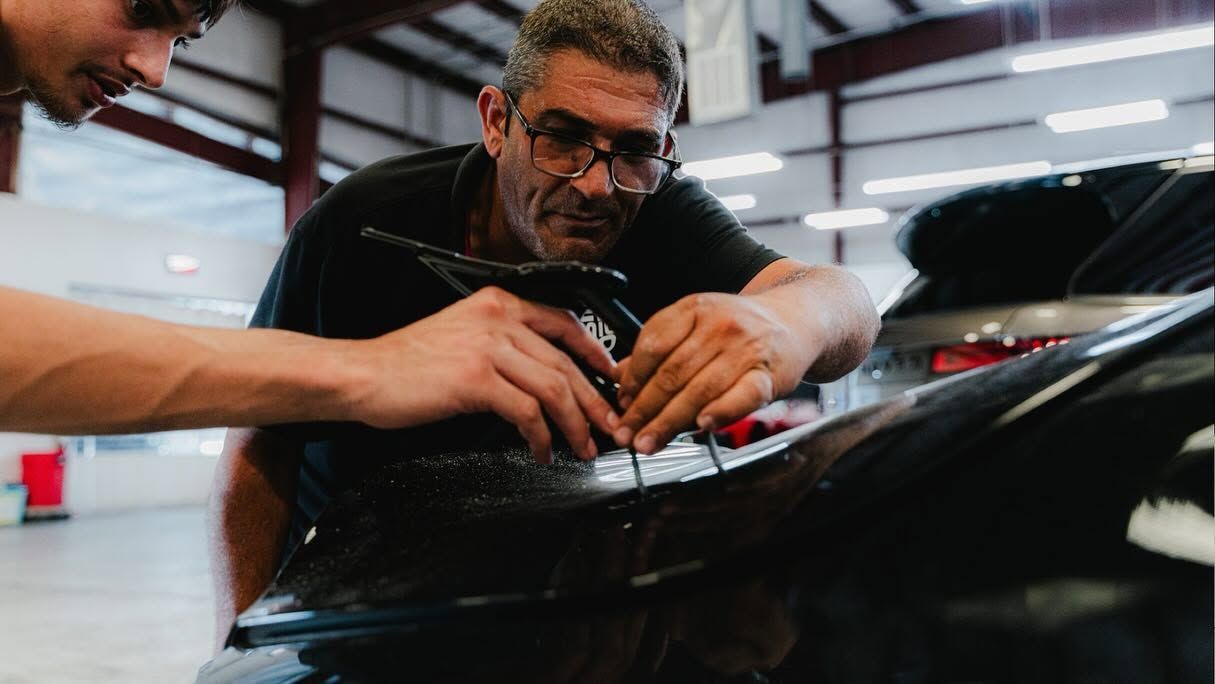 Two people work on a glossy black car in a garage, using a tool near the hood.