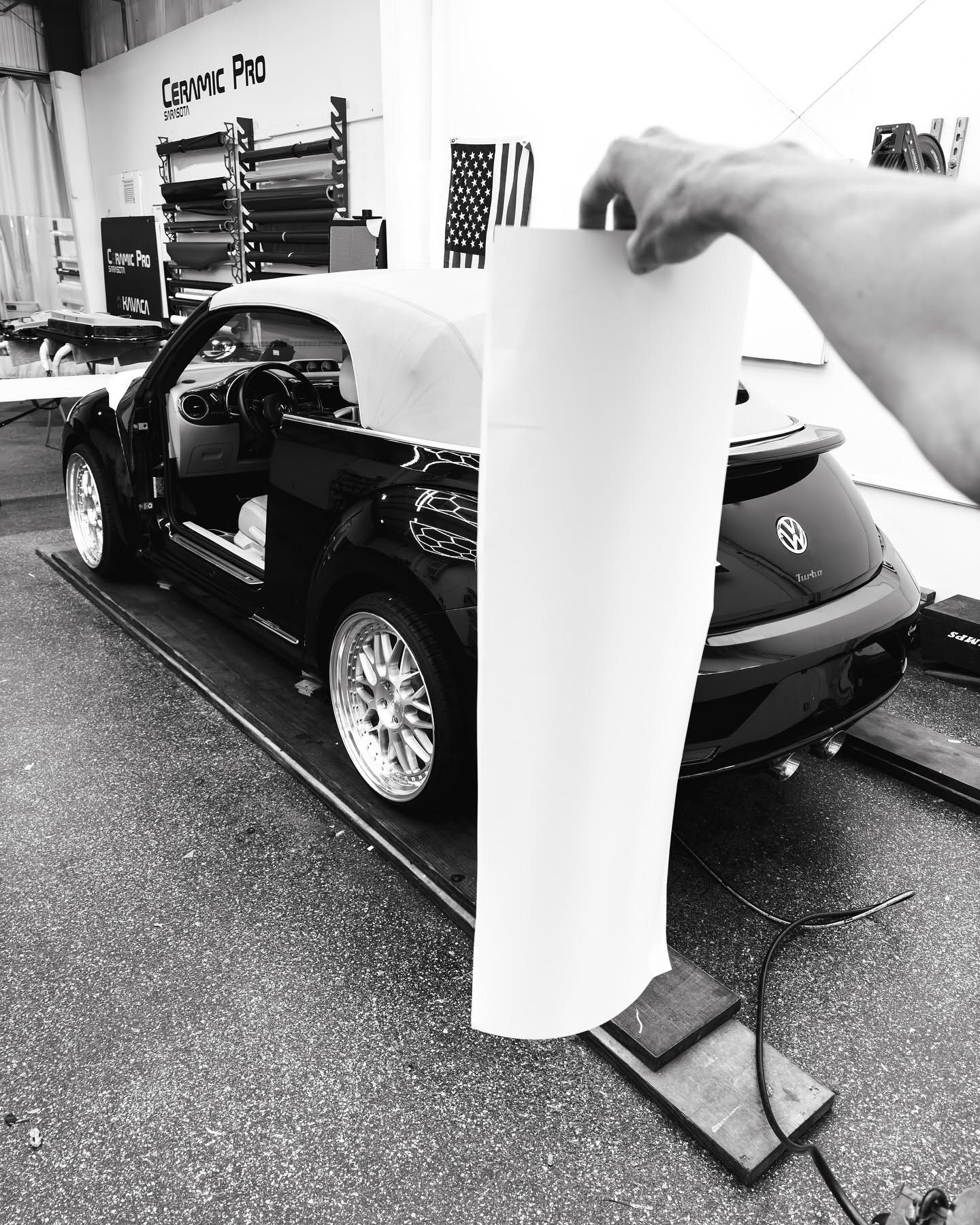 Black car on a lift in a garage, with a person holding a tall white panel beside it.