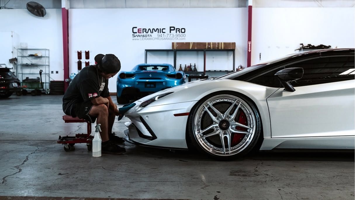 Mechanic working on a white sports car in a garage with a “Ceramic Pro” sign behind it