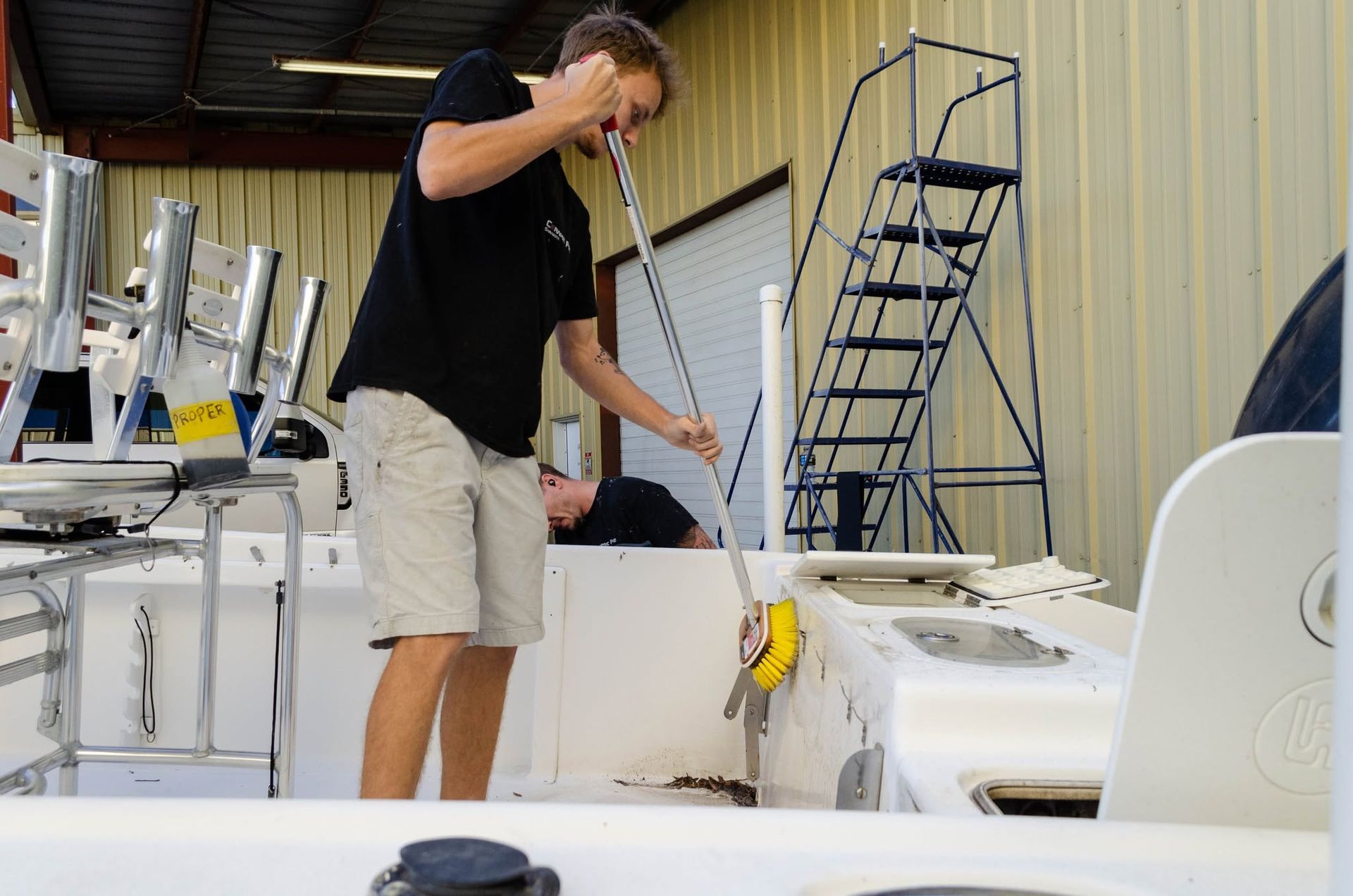 Man in black shirt and khaki shorts working on a boat in a storage area, with ladders and equipment nearby
