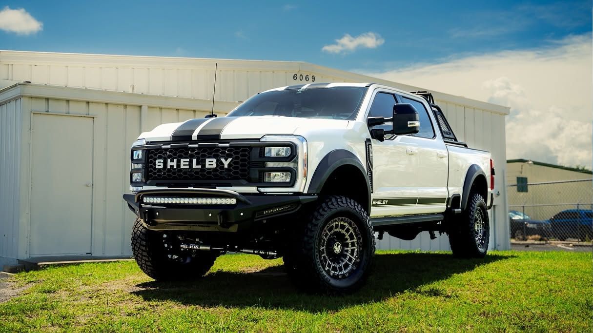 White Shelby pickup truck parked on grass beside white buildings under a bright blue sky