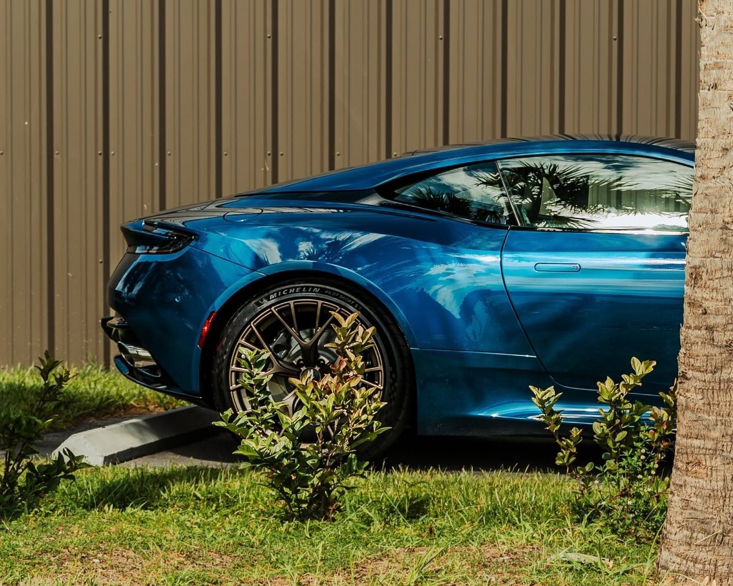Blue sports car parked on grass beside a tan fence, front wheel near a curb.
