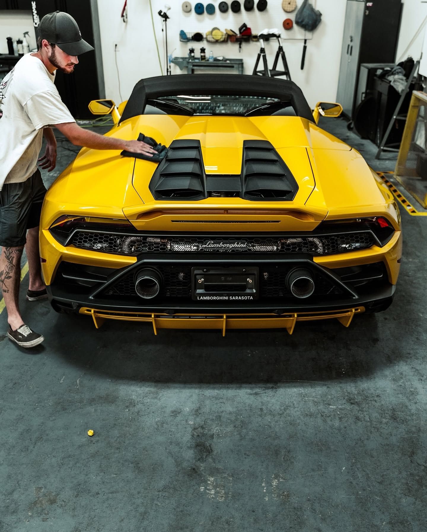 Man in garage polishing a yellow sports car with black accents and rear engine vents