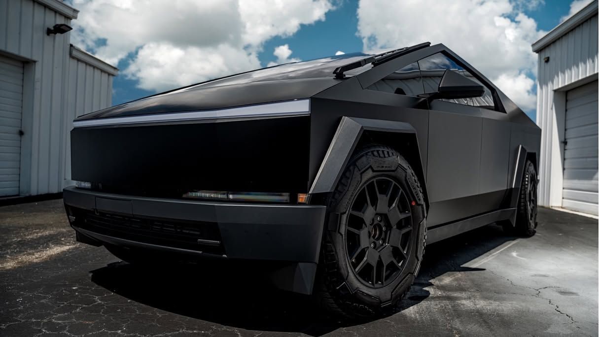 Matte black futuristic pickup truck parked between white industrial buildings under cloudy sky