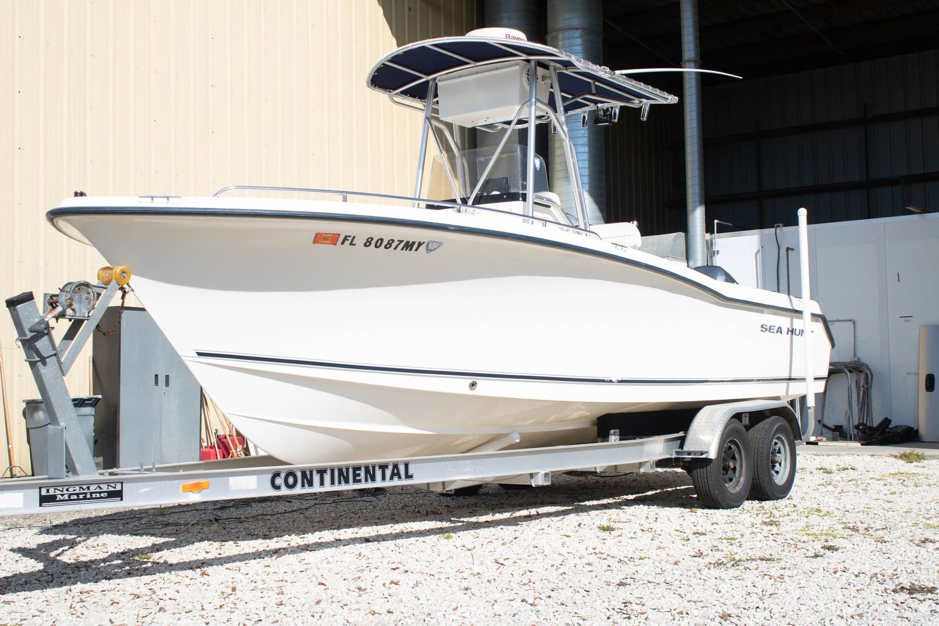 White center-console boat on a trailer outside a marina building, with a T-top and blue cover.