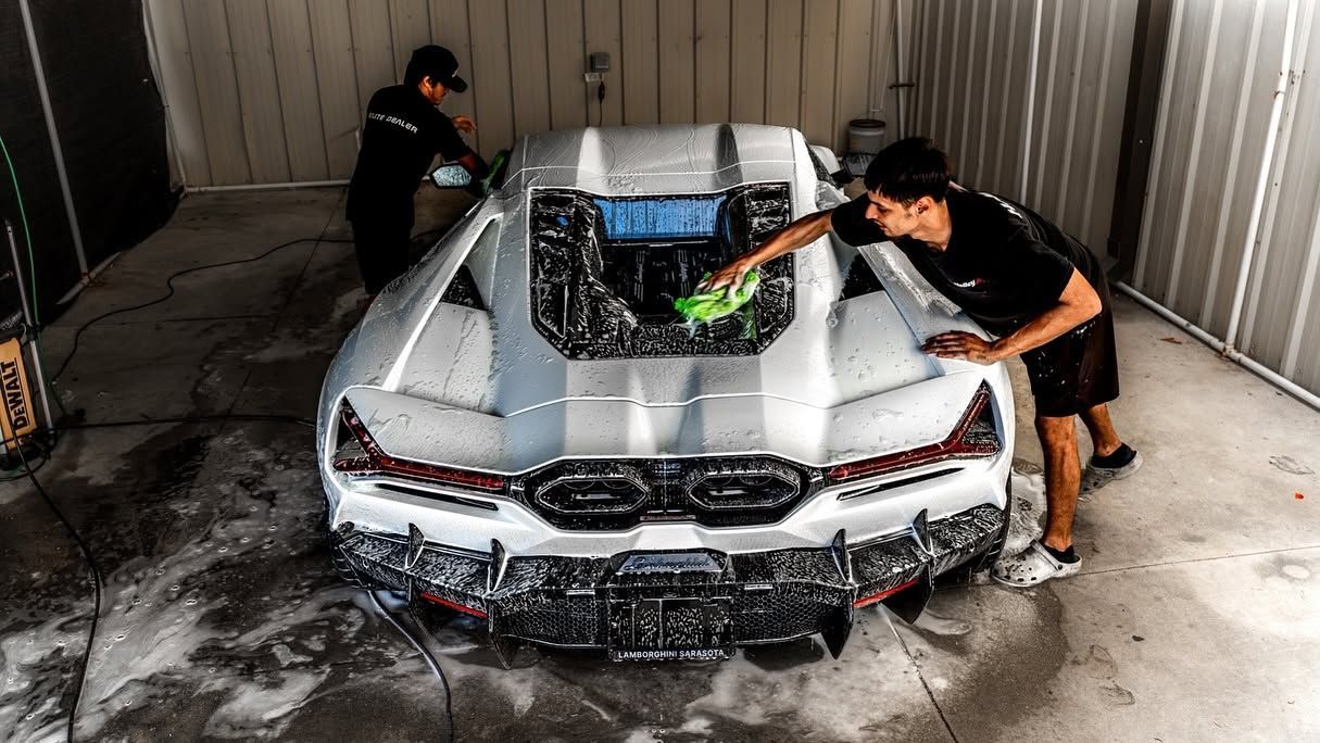 Two people washing a silver sports car in a garage, with water splashing over the hood.