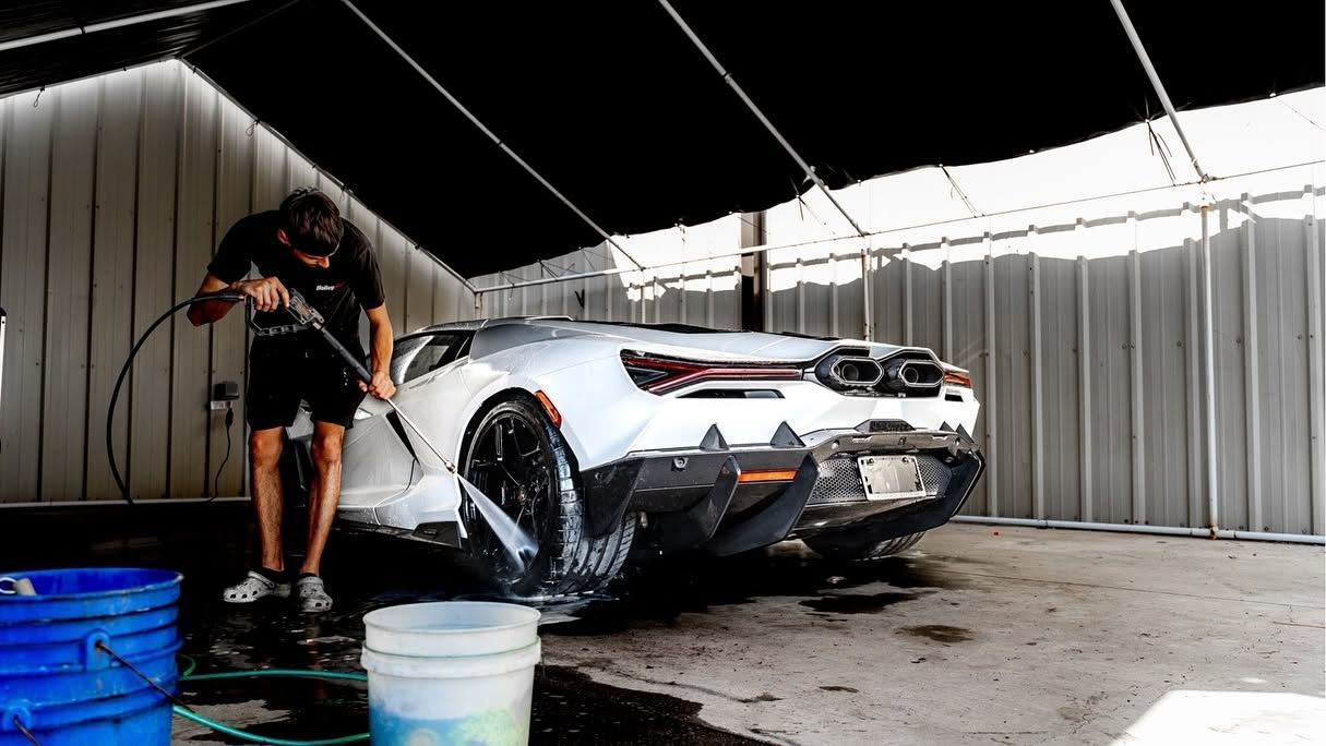 Man washing a white sports car in a covered car wash bay, with buckets and water on the floor