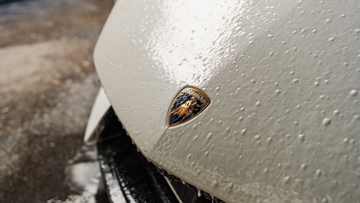 Close-up of a white sports car hood with a gold emblem and water droplets on the surface