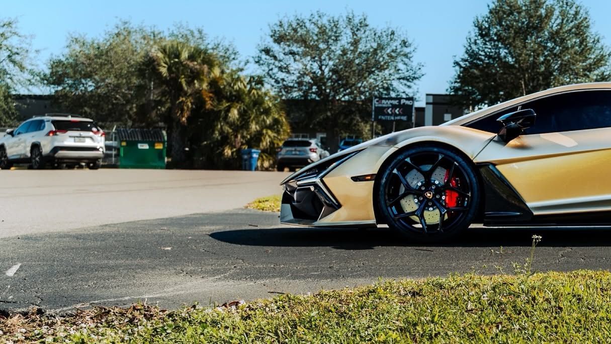 Low-angle view of a gold sports car parked by a road, with palm trees and other cars in the background.