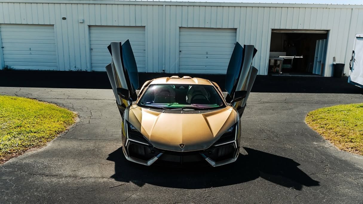 Gold sports car with doors raised, parked on a driveway in front of a white garage building