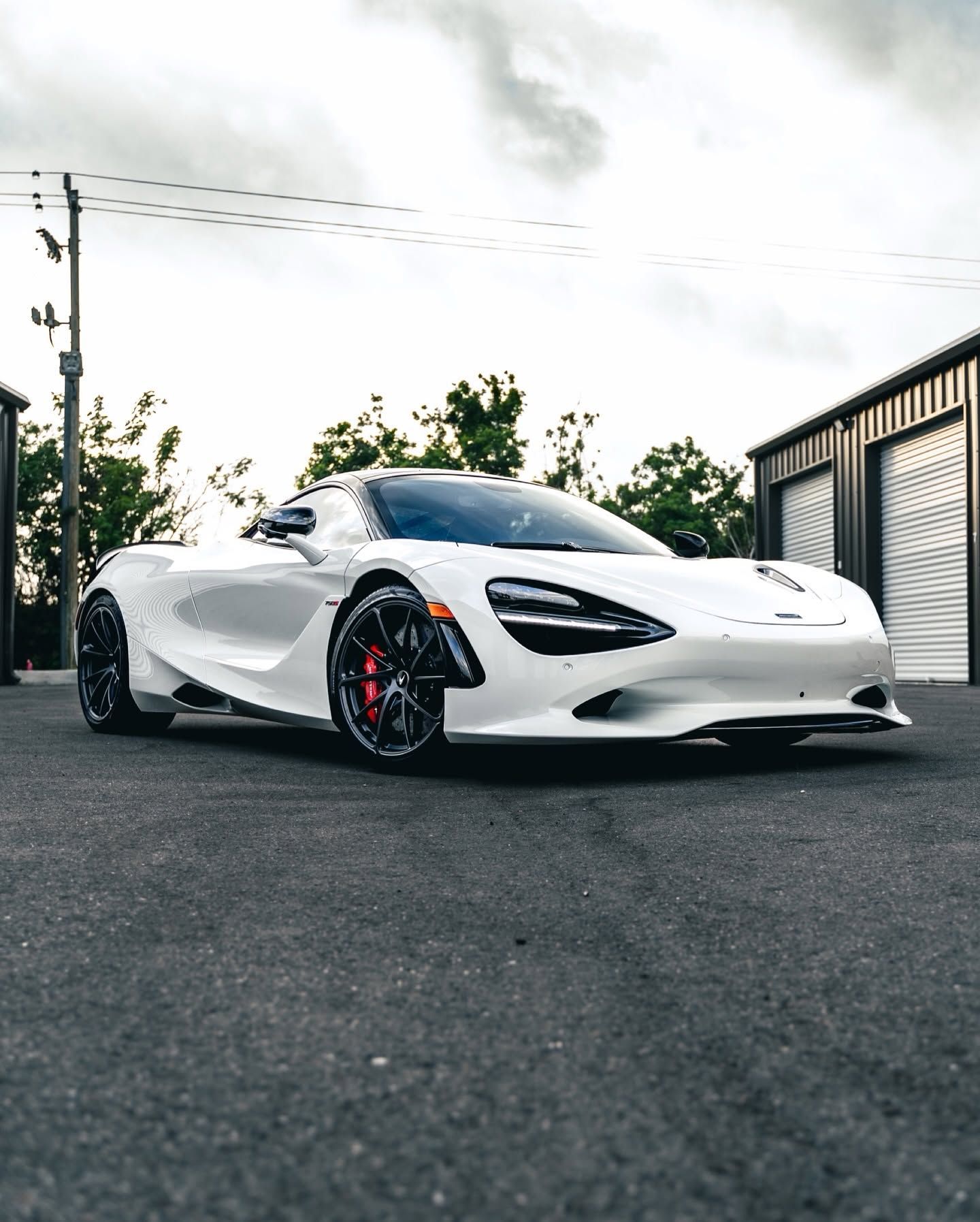 White sports car parked on asphalt beside a garage, with red brake calipers and a cloudy sky.