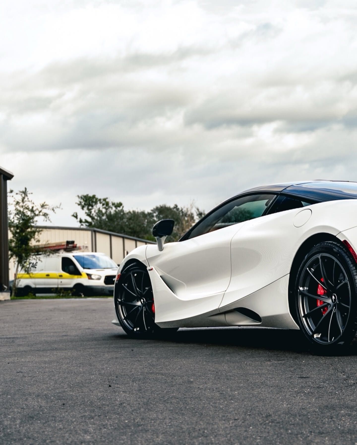 White sports car parked on asphalt, with a yellow car and cloudy sky in the background