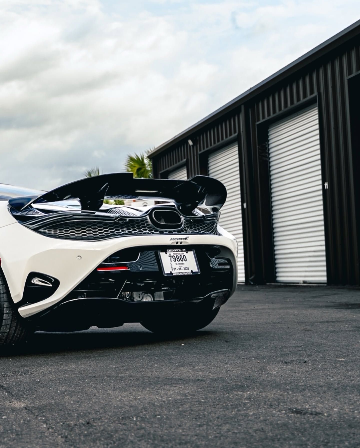 White sports car with rear hatch open, parked beside black garage doors on a cloudy day