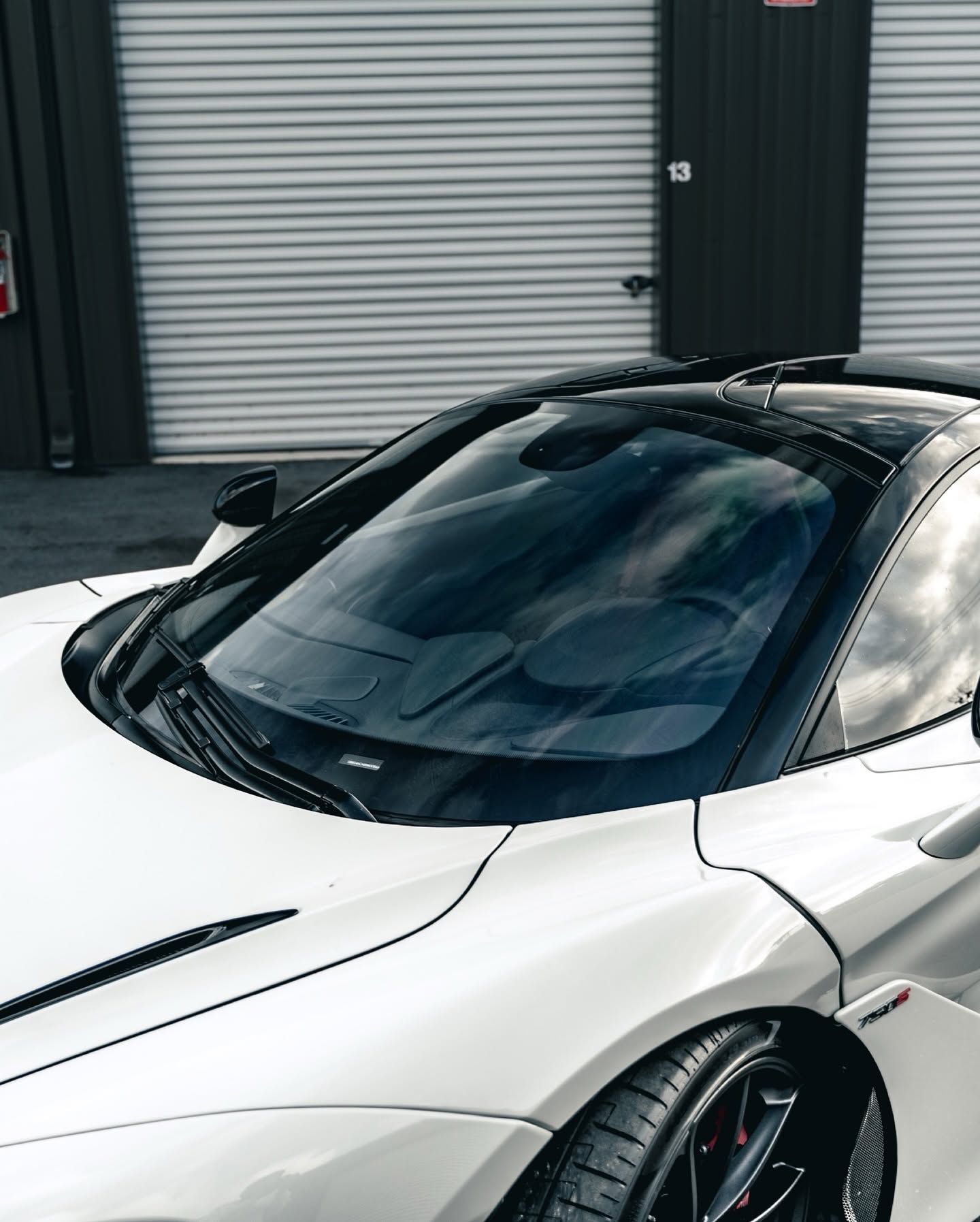 White sports car parked in front of a garage, with a sleek black roof and windshield reflection.