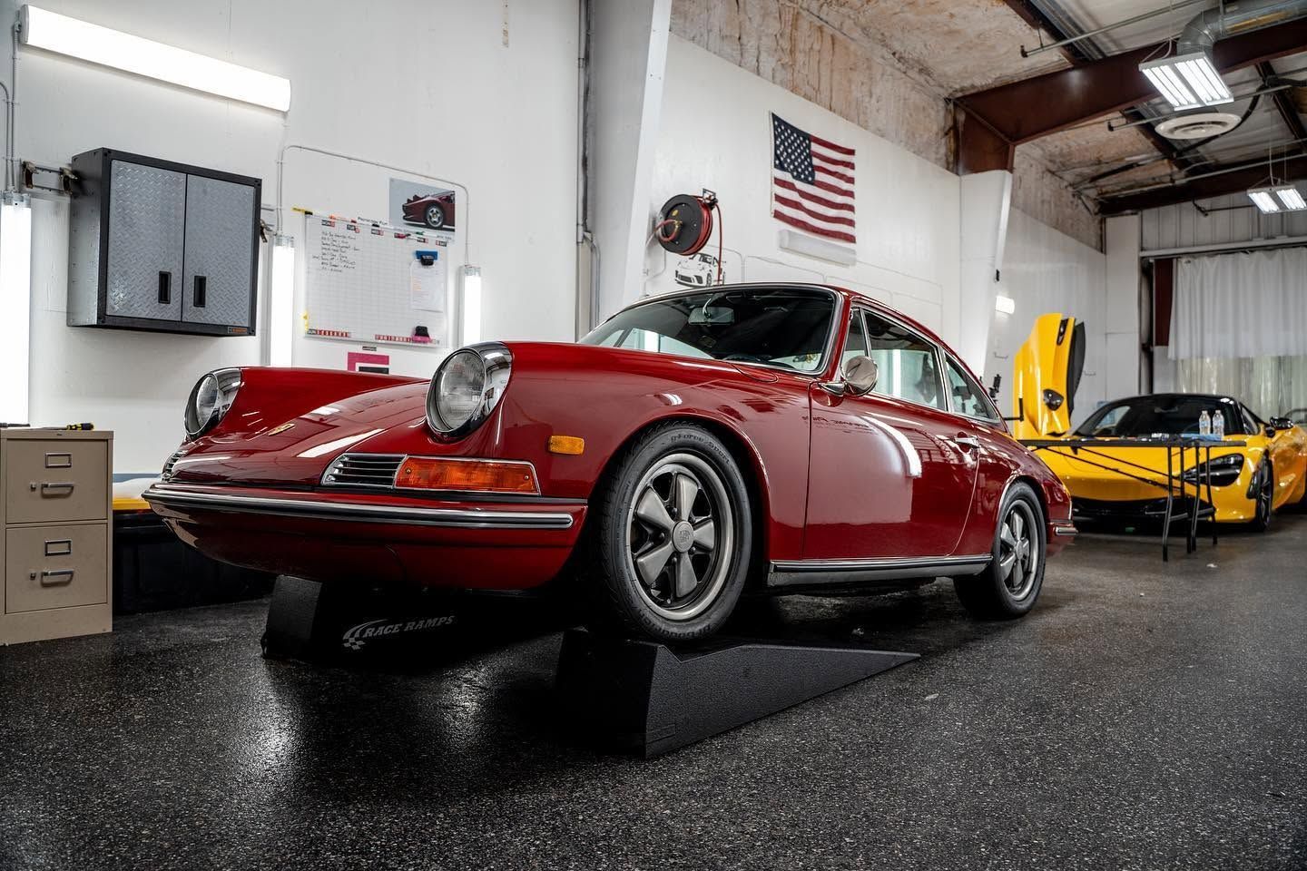 Red vintage Porsche in a showroom beside a yellow sports car, with an American flag on the wall.
