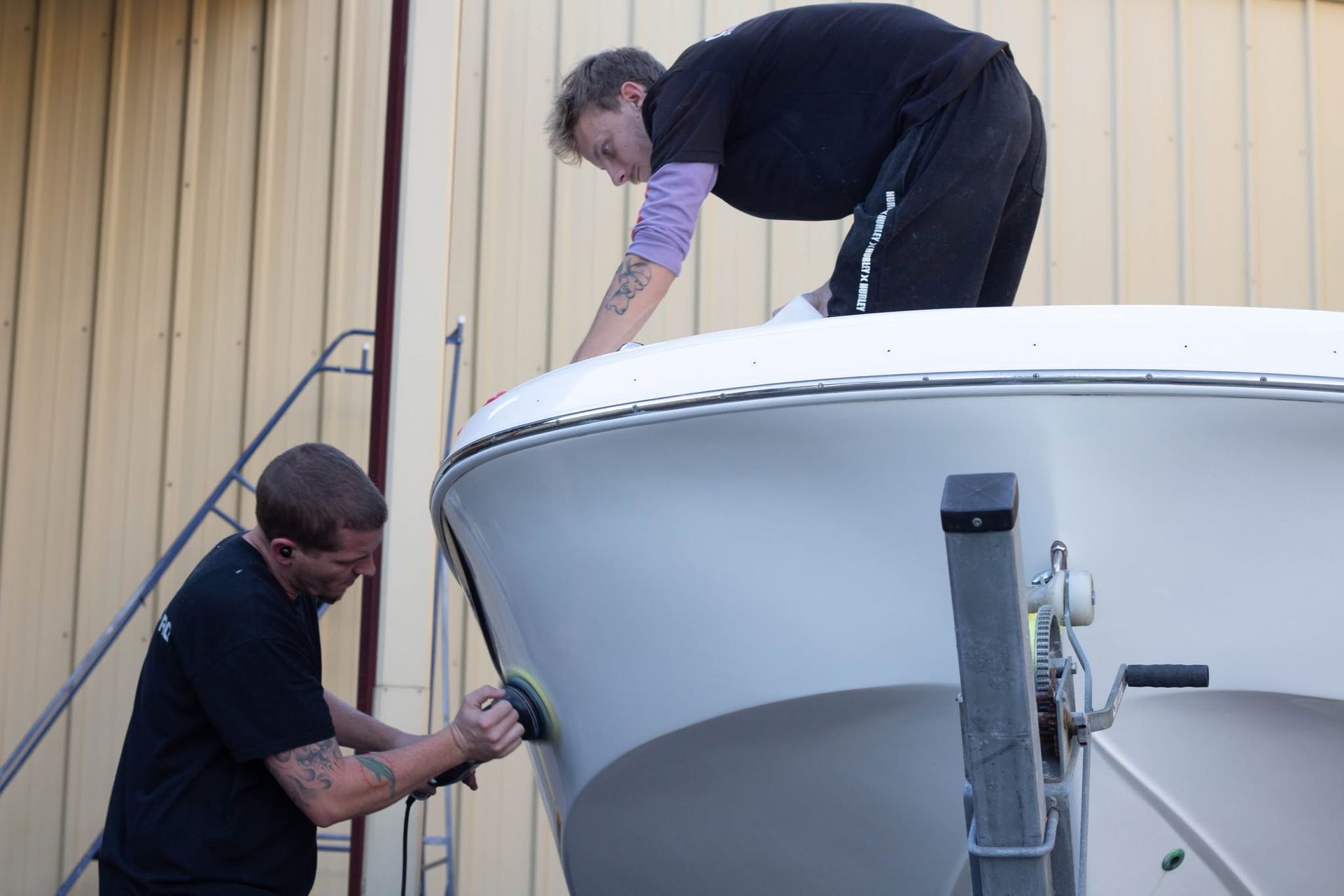 Two people working on a white boat beside a tan building, one polishing the hull.